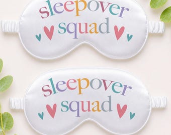 May include: Two white sleep masks with the words "sleepover squad" in colorful letters. Each mask has a pink heart and two smaller teal hearts. The masks have elastic bands for a comfortable fit.