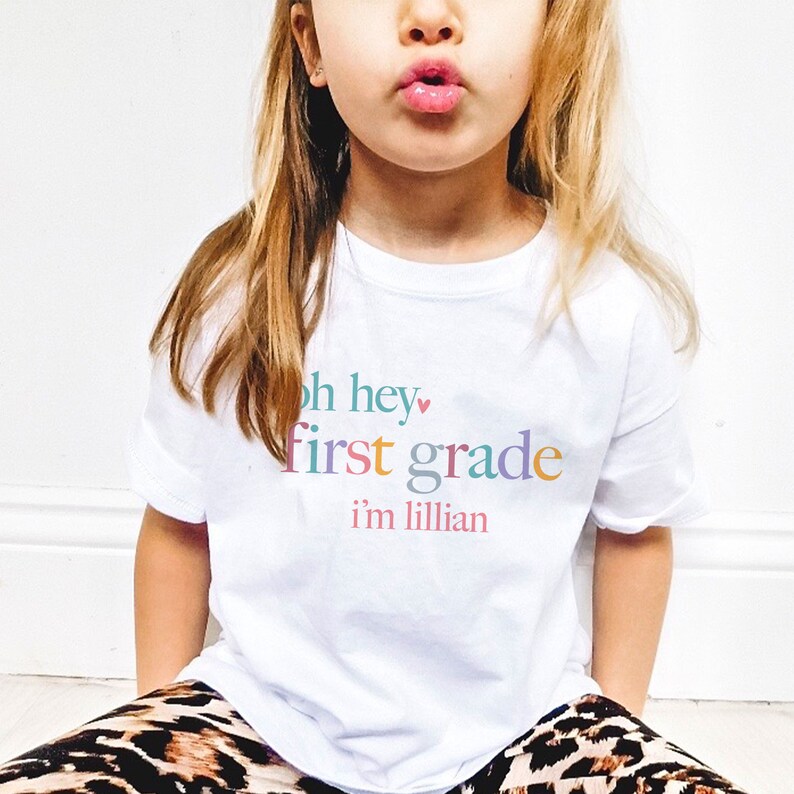 May include: A white t-shirt with the text "oh hey first grade i'm lillian" in colorful letters. The text is in a playful font and includes a small pink heart.