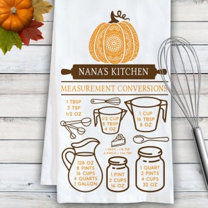 May include: A white kitchen towel featuring a brown and orange graphic of a pumpkin, a rolling pin with the text NANA'S KITCHEN, and a measurement conversion chart. The chart includes illustrations of measuring cups, spoons, and jars with text.
