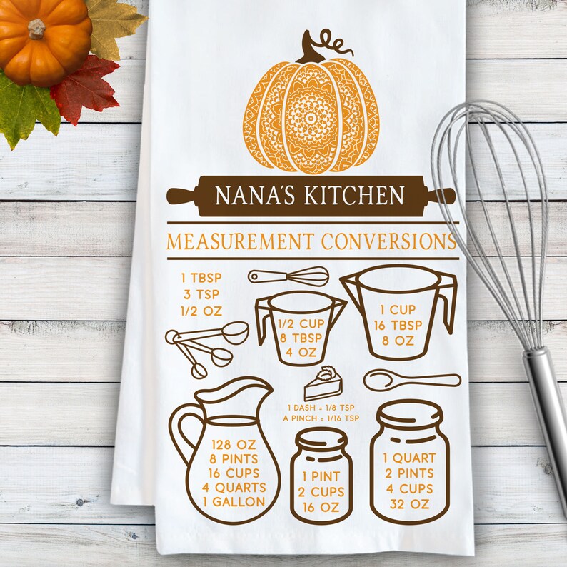 May include: A white kitchen towel featuring a brown and orange graphic of a pumpkin, a rolling pin with the text NANA'S KITCHEN, and a measurement conversion chart. The chart includes illustrations of measuring cups, spoons, and jars with text.
