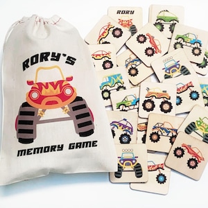 May include: A cream-colored drawstring fabric bag with the text RORY'S MEMORY GAME and a red monster truck graphic, accompanied by a collection of square wooden tiles featuring various colorful monster truck illustrations for a matching game.