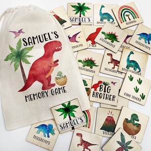 May include: A beige drawstring fabric bag labeled Samuel's Memory Game with a red dinosaur and palm tree illustration, accompanied by numerous square wooden tiles featuring watercolor dinosaur images, volcano, rainbow, and text labels.