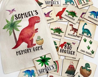 May include: A beige drawstring fabric bag labeled Samuel's Memory Game with a red dinosaur and palm tree illustration, accompanied by numerous square wooden tiles featuring watercolor dinosaur images, volcano, rainbow, and text labels.