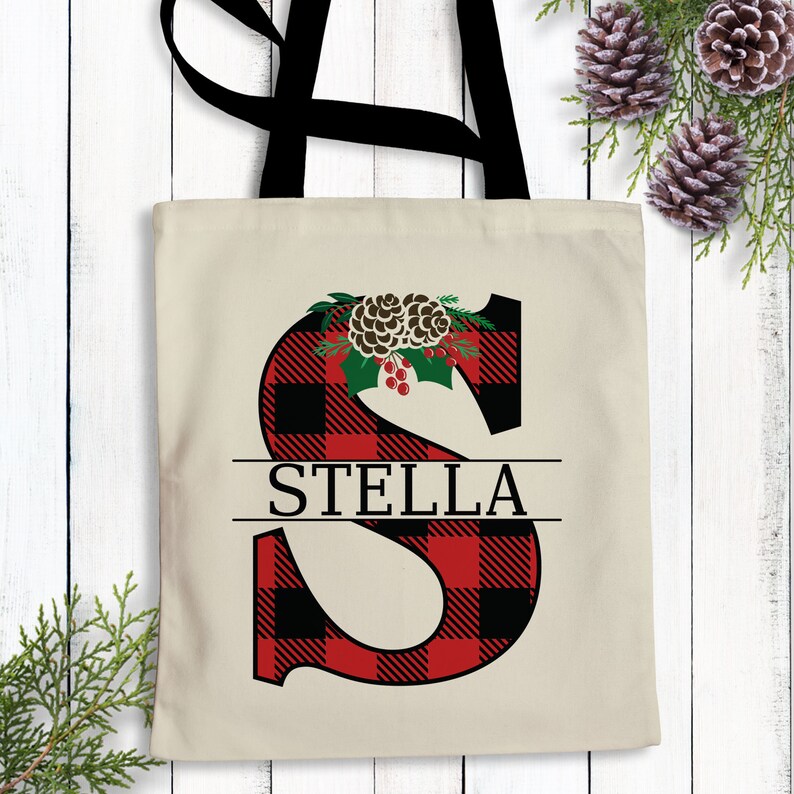 Pu&ograve; includere: Una borsa tote in tela di cotone rossa e nera a quadri di bufalo con un manico nero. La borsa presenta una grande lettera "S" con un designo di pigna e bacche di agrifoglio. Il nome "STELLA" &egrave; stampato in nero sotto la lettera.