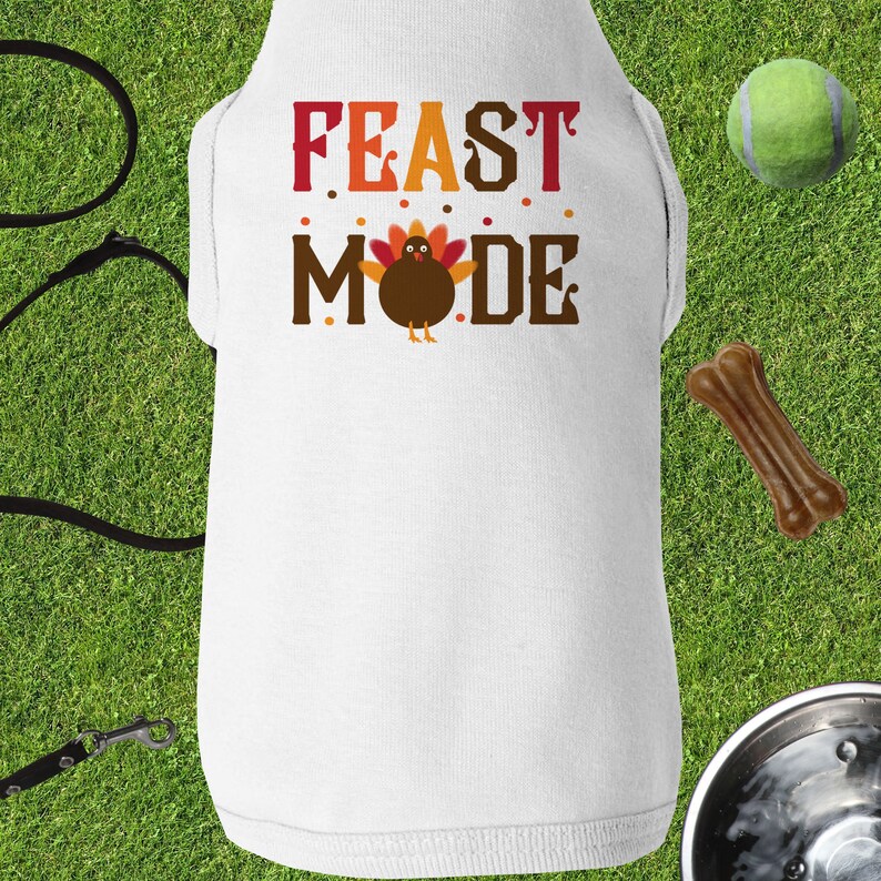 Op de afbeelding: Witte hondenvest met de tekst "FEAST MODE" in rode, oranje en bruine letters. Een cartoon-kalkoen bevindt zich in het midden van de tekst.