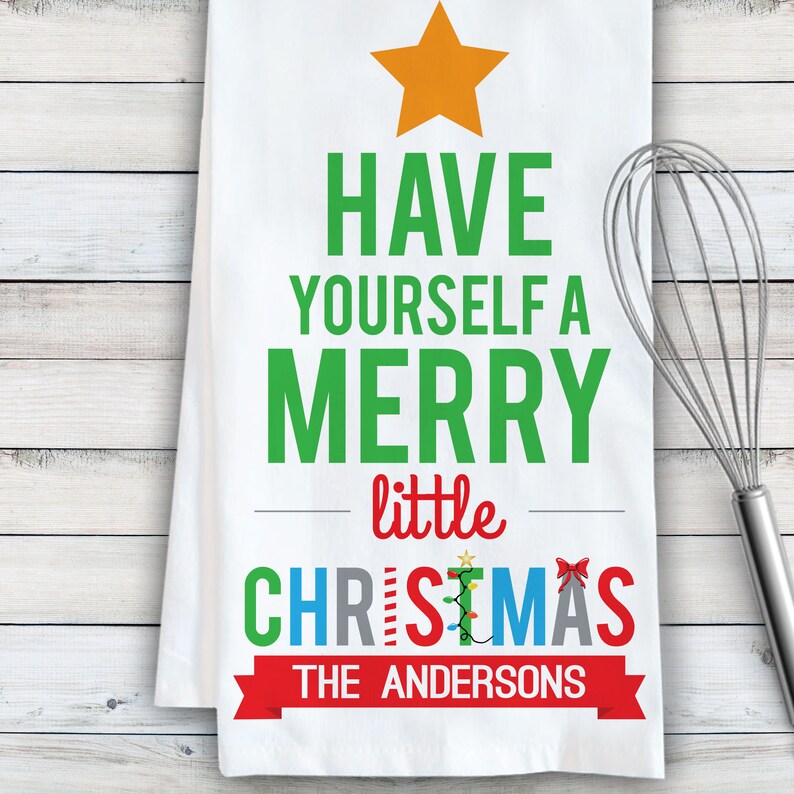 May include: White kitchen towel with a green Christmas tree design. The tree has a star on top and the text "Have yourself a merry little Christmas" with the name "The Andersons" at the bottom.