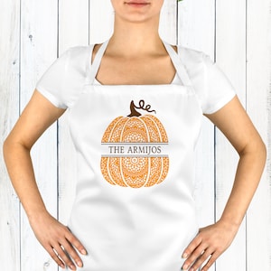 May include: A white apron featuring a central orange pumpkin graphic with a detailed mandala pattern. The pumpkin is split horizontally by a line containing the text THE ARMIJOS in dark brown lettering, topped with a brown stem and vine.
