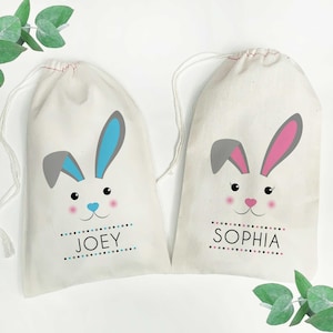 May include: Two white cotton drawstring bags with bunny faces. The bag on the left has a blue bunny with the name "JOEY" printed below. The bag on the right has a pink bunny with the name "SOPHIA" printed below.