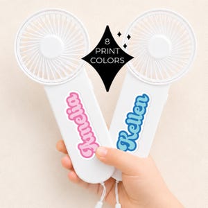 May include: Two white handheld fans with a circular fan head and a handle. One fan has the name "Amelia" in pink, and the other has "Kellen" in blue. A black diamond shape with the text "8 PRINT COLORS" is in the center.
