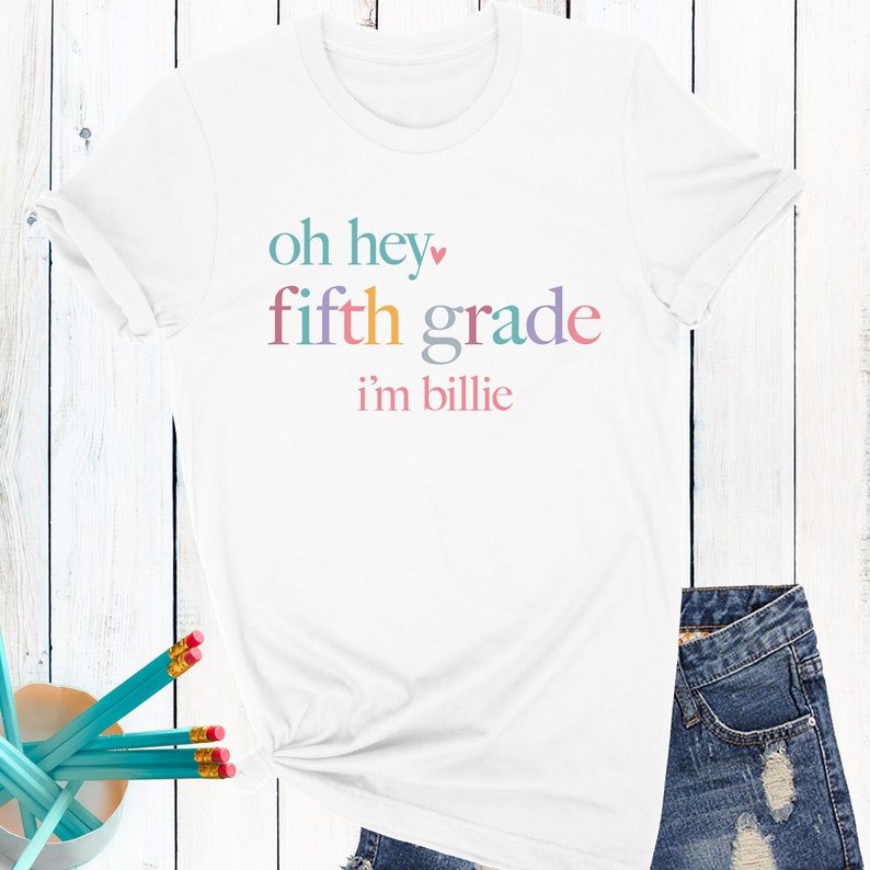 May include: White t-shirt with the text "oh hey fifth grade i'm billie" in rainbow colors.