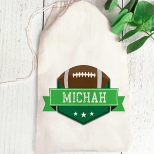 May include: A white drawstring bag with a green and black football-themed design. The design features a brown football with white laces, a green banner with the name "MICHAH" in white text, and three white stars.