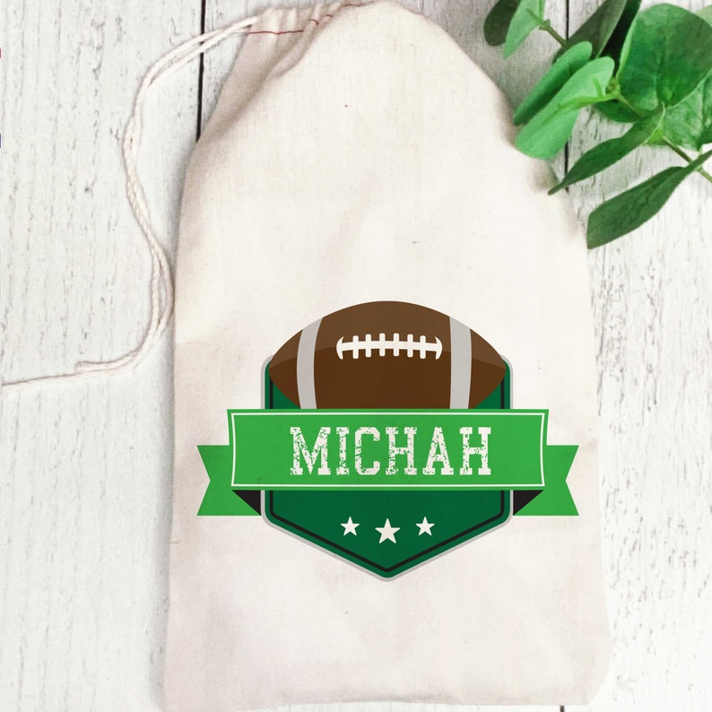 May include: A white drawstring bag with a green and black football-themed design. The design features a brown football with white laces, a green banner with the name "MICHAH" in white text, and three white stars.