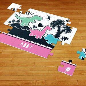 May include: A jigsaw puzzle featuring a white background with black, mint green, pink, and teal dinosaur illustrations, palm trees, and the text ROAR!!! and ABBY. The puzzle pieces are scattered, showing a mix of completed sections and loose pieces.