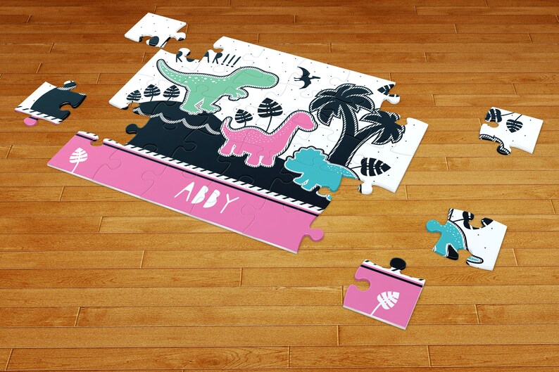 May include: A jigsaw puzzle featuring a white background with black, mint green, pink, and teal dinosaur illustrations, palm trees, and the text ROAR!!! and ABBY. The puzzle pieces are scattered, showing a mix of completed sections and loose pieces.