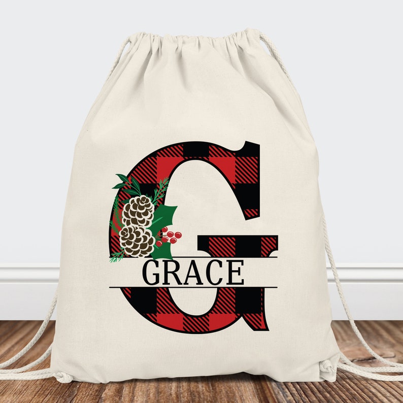 Pu&ograve; includere: Una borsa a cordoncino bianca con una lettera G a quadri rossi e neri e il nome "GRACE" stampato sotto. La lettera G &egrave; decorata con una pigna e un ramo di agrifoglio.
