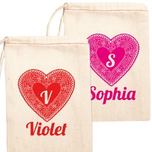 May include: Two white cotton drawstring bags with red and pink lace hearts. The hearts have the letters V and S in the center. The bags are personalized with the names Violet and Sophia.