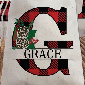 Pu&ograve; includere: Una lettera G in tessuto a quadri rosso e nero con un motivo di pigna e ramo di agrifoglio. Il nome "GRACE" &egrave; scritto in lettere nere sotto la lettera.
