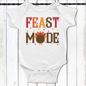 K&ouml;nnte beinhalten: Wei&szlig;er Baby-Strampler mit dem Text "FEAST MODE" in braunen, orangen und roten Buchstaben. Ein Cartoon-Truthahn mit orangefarbenen, roten und braunen Federn befindet sich in der Mitte des Textes.