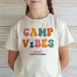 May include: A light beige t-shirt with the text "CAMP VIBES" in colorful, retro-style letters. The text "RILEY CAMP MANITOQUA" is printed below in black.