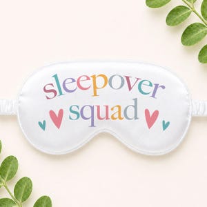 Puede incluir: M&aacute;scara para dormir blanca con las palabras "sleepover squad" en letras coloridas. La m&aacute;scara tiene un dise&ntilde;o de coraz&oacute;n y una banda el&aacute;stica. La m&aacute;scara para dormir est&aacute; dise&ntilde;ada para bloquear la luz y promover la relajaci&oacute;n.