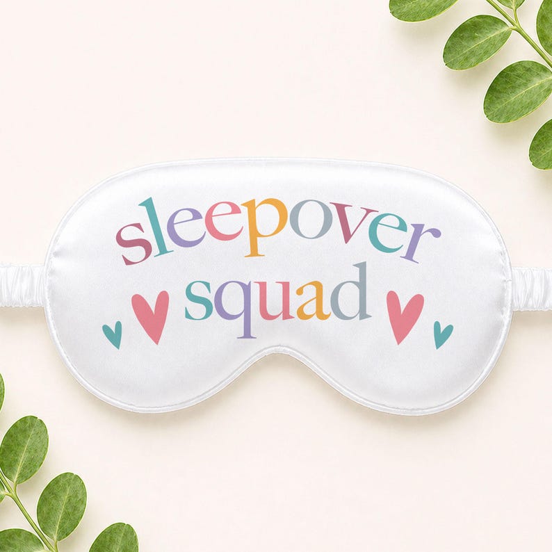 Puede incluir: M&aacute;scara para dormir blanca con las palabras "sleepover squad" en letras coloridas. La m&aacute;scara tiene un dise&ntilde;o de coraz&oacute;n y una banda el&aacute;stica. La m&aacute;scara para dormir est&aacute; dise&ntilde;ada para bloquear la luz y promover la relajaci&oacute;n.