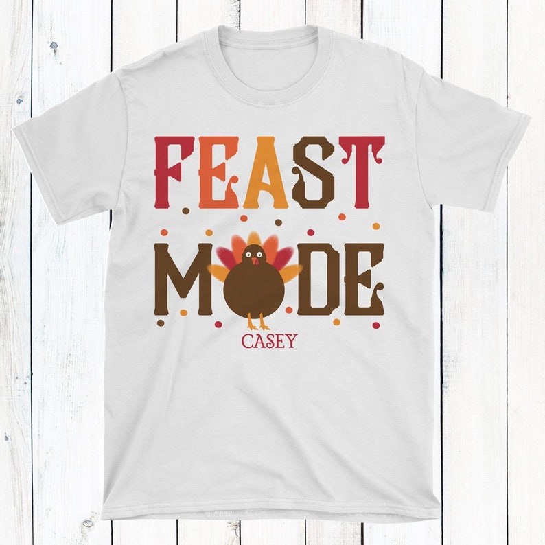 K&ouml;nnte beinhalten: Wei&szlig;es T-Shirt mit dem Text "FEAST MODE" in roten, orangen und braunen Buchstaben. Ein Cartoon-Truthahn befindet sich in der Mitte des Wortes "MODE". Der Name "CASEY" ist unter dem Truthahn gedruckt.
