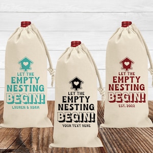 May include: Three beige wine bottle bags with black drawstring closures. Each bag has a black and white or red and white graphic of a birdhouse with a heart on it and the text "Let the Empty Nesting Begin!" with personalized text below. The bags are on a wood surface.