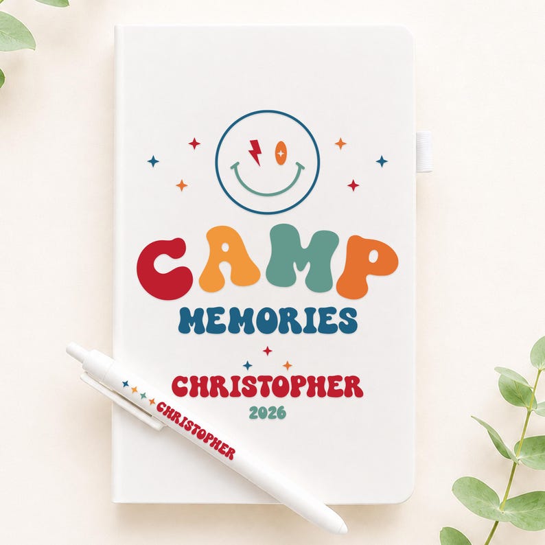 May include: A white notebook featuring a colorful graphic of a smiley face with a lightning bolt eye, the text CAMP MEMORIES, the name CHRISTOPHER, and the year 2026. A matching white pen with the name CHRISTOPHER printed on it rests across the cover.