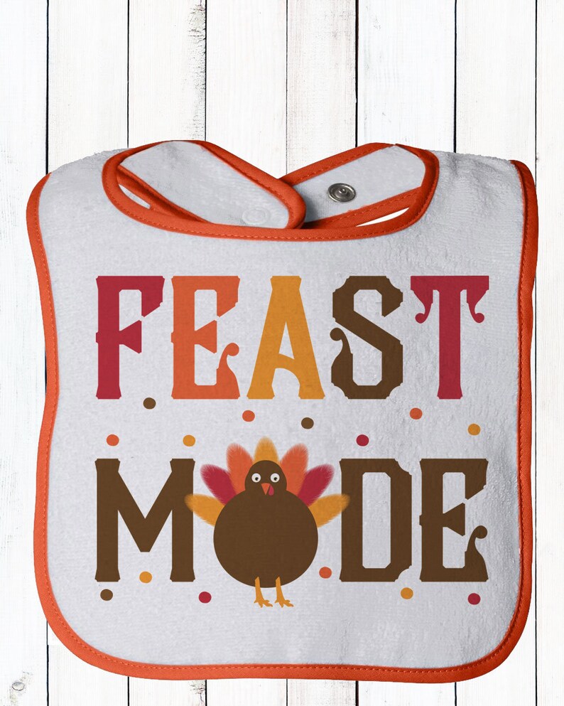 K&ouml;nnte beinhalten: Wei&szlig;er Baby-L&auml;tzchen mit orangefarbenem Rand. Das L&auml;tzchen zeigt einen braunen Truthahn mit orangefarbenen und roten Federn und dem Text "FEAST MODE" in Orange, Rot und Braun.