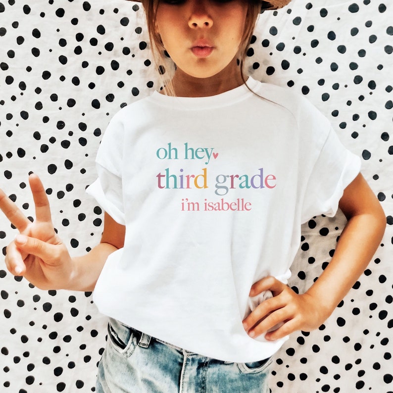 May include: White t-shirt with the text "oh hey third grade i'm isabelle" in rainbow colors. The text includes a small pink heart.