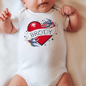 May include: White baby onesie with a red heart and two blue birds. The heart has a banner with the name "Brody" on it.