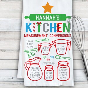 May include: A white kitchen tea towel with a red, green, and blue "KITCHEN" design with Christmas lights and a star. The towel features a list of kitchen measurement conversions in ounces, cups, pints, quarts, and gallons.
