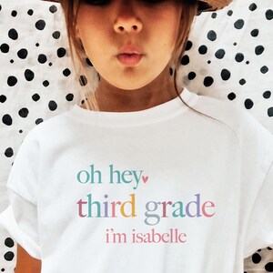 May include: White t-shirt with the text "oh hey third grade i'm isabelle" in colorful letters.