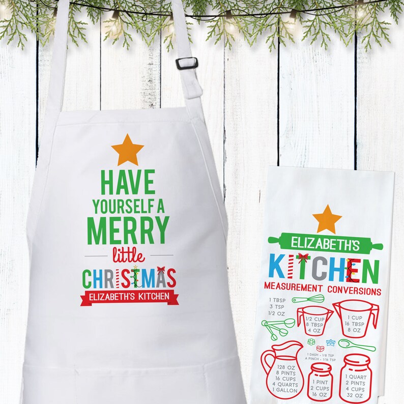 May include: A white kitchen apron with a green and red Christmas tree design. The text reads "Have yourself a merry little Christmas, Elizabeth's Kitchen". A white tea towel with a red and blue rolling pin design and the text "Elizabeth's Kitchen" is also shown. The tea towel has a chart of kitchen measurement conversions.