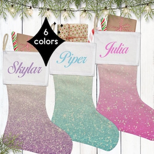 May include: Three personalized Christmas stockings with ombre glitter fabric. The stockings are pink, teal, and purple. The names "Skylar", "Piper", and "Julia" are embroidered on the white cuff of each stocking.