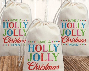 May include: Three off-white drawstring canvas bags featuring the text HAVE A HOLLY JOLLY Christmas in colorful red, blue, green, and pink lettering. Each bag is personalized with a name at the bottom, specifically Henry, Ingrid, and Parker.