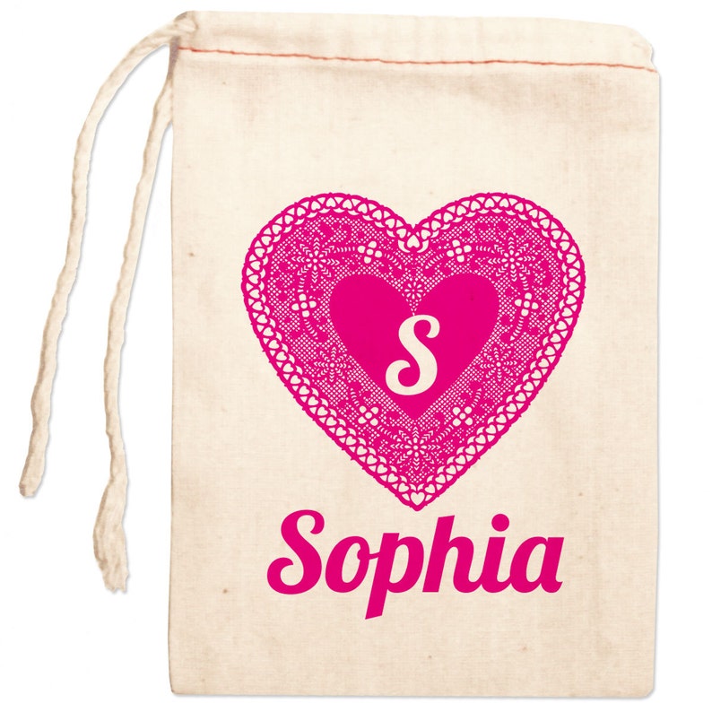 May include: A white cotton drawstring bag with a pink heart design. The heart has a pink lace pattern and a white letter "S" in the center. The name "Sophia" is printed below the heart in pink.