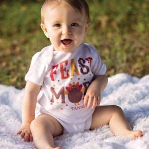 K&ouml;nnte beinhalten: Ein Baby tr&auml;gt einen wei&szlig;en Strampler mit dem Text "FEAST MODE" und einer Cartoon-Truthahn-Grafik. Der Strampler hat eine braune und orangefarbene Farbgebung. Das Baby sitzt auf einer wei&szlig;en, flauschigen Oberfl&auml;che.