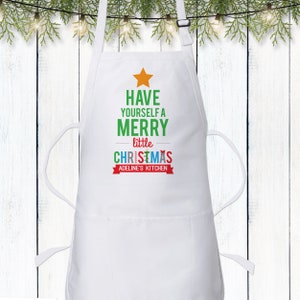 May include: White kitchen apron with a green Christmas tree design. The tree has a yellow star on top and the text "Have yourself a merry little Christmas" in red, green, blue, and yellow. The text "Adeline's Kitchen" is in red on a white banner.