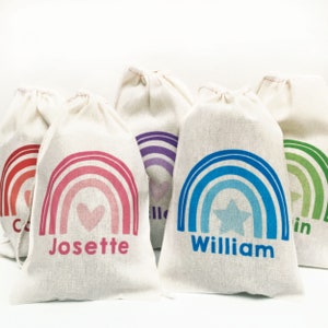 May include: Five personalized rainbow-themed gift bags with different colors and designs. The bags are made of natural fabric and have a drawstring closure. The names "William", "Josette", "Colin", "Ella", and "Erin" are printed on the bags.