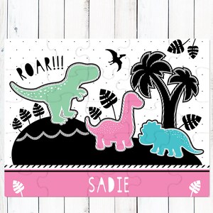May include: A rectangular jigsaw puzzle featuring a black, white, mint green, and pink dinosaur theme. The design includes a mint T-Rex with the text ROAR!!!, a pink long-necked dinosaur, a blue triceratops, palm trees, and the name SADIE.