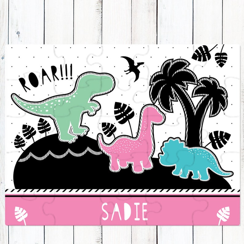 May include: A rectangular jigsaw puzzle featuring a black, white, mint green, and pink dinosaur theme. The design includes a mint T-Rex with the text ROAR!!!, a pink long-necked dinosaur, a blue triceratops, palm trees, and the name SADIE.