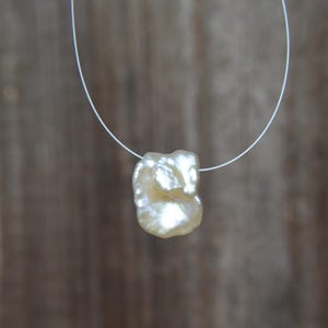 May include: A single, irregular, off-white baroque pearl pendant suspended on a thin, clear nylon cord. The pearl features a lustrous, textured surface with organic, asymmetrical curves and a high-shine finish that reflects light across its surface.