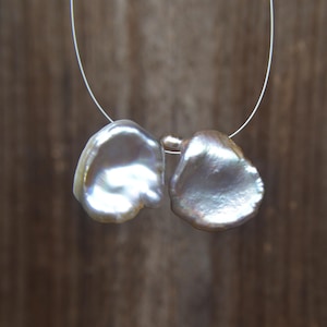 May include: Two irregular, lustrous, off-white baroque pearls are strung together on a thin, clear filament. The pearls exhibit a smooth, iridescent surface with subtle pink and cream undertones, showcasing a unique, organic, and flattened shape.