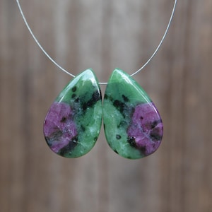 May include: Two teardrop-shaped gemstones. Each gem features a vibrant green base with dark spots and a prominent magenta-colored center. The stones are strung on a thin, silver-colored wire, suggesting they are intended for jewelry making.