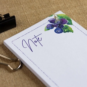 May include: A white notepad with a watercolor illustration of blueberries and green leaves. The word "Note" is written in cursive in the upper left corner. A black binder clip is visible in the upper left corner.