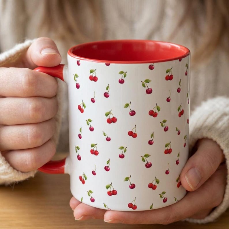 May include: A white ceramic mug featuring a repeating pattern of small red cherries with green stems and leaves. The mug has a bright red handle and a solid red interior, providing a vibrant contrast to the white exterior surface.