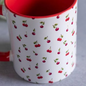 May include: A white ceramic mug featuring a repeating pattern of small red cherries with green stems and leaves. The interior of the mug and the handle are finished in a solid, vibrant red colour, providing a contrast to the white exterior surface.