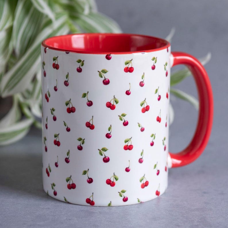 May include: A white ceramic mug featuring a repeating pattern of small red cherries with green stems and leaves. The interior of the mug and the handle are a solid, vibrant red colour. The mug has a cylindrical shape with a smooth, glossy finish.
