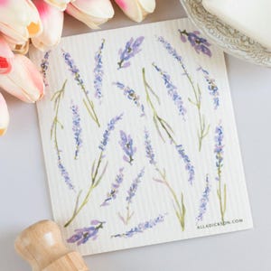 May include: A white, square cleaning cloth featuring a watercolor design of lavender sprigs in shades of purple and green. The cloth has a textured appearance and the text "ALLADICKSON.COM" is visible in the lower right corner. The cloth is surrounded by other objects.